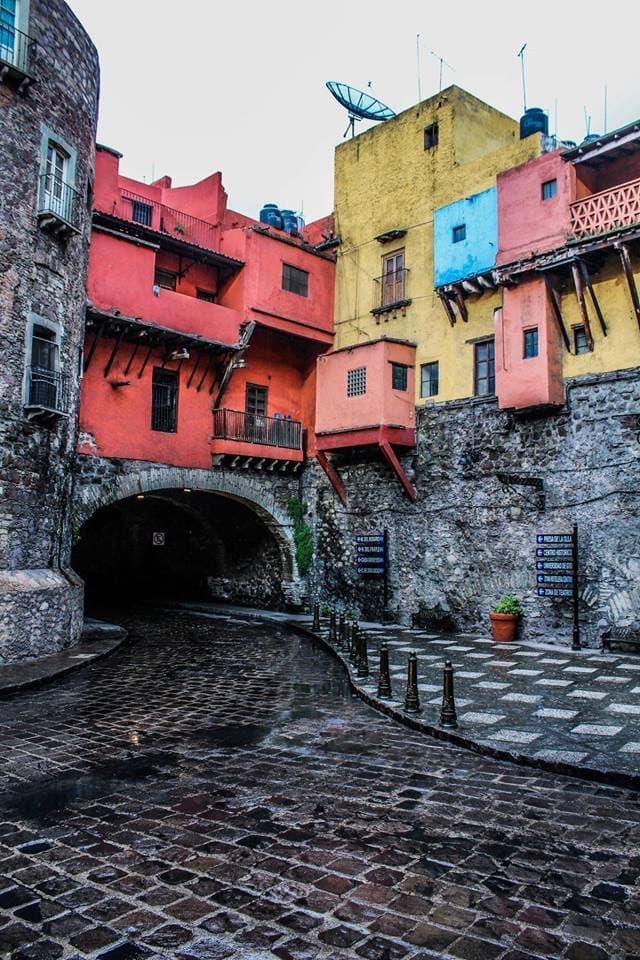 A wet cobbled street curving into a stone tunnel, under red, yellow and blue houses